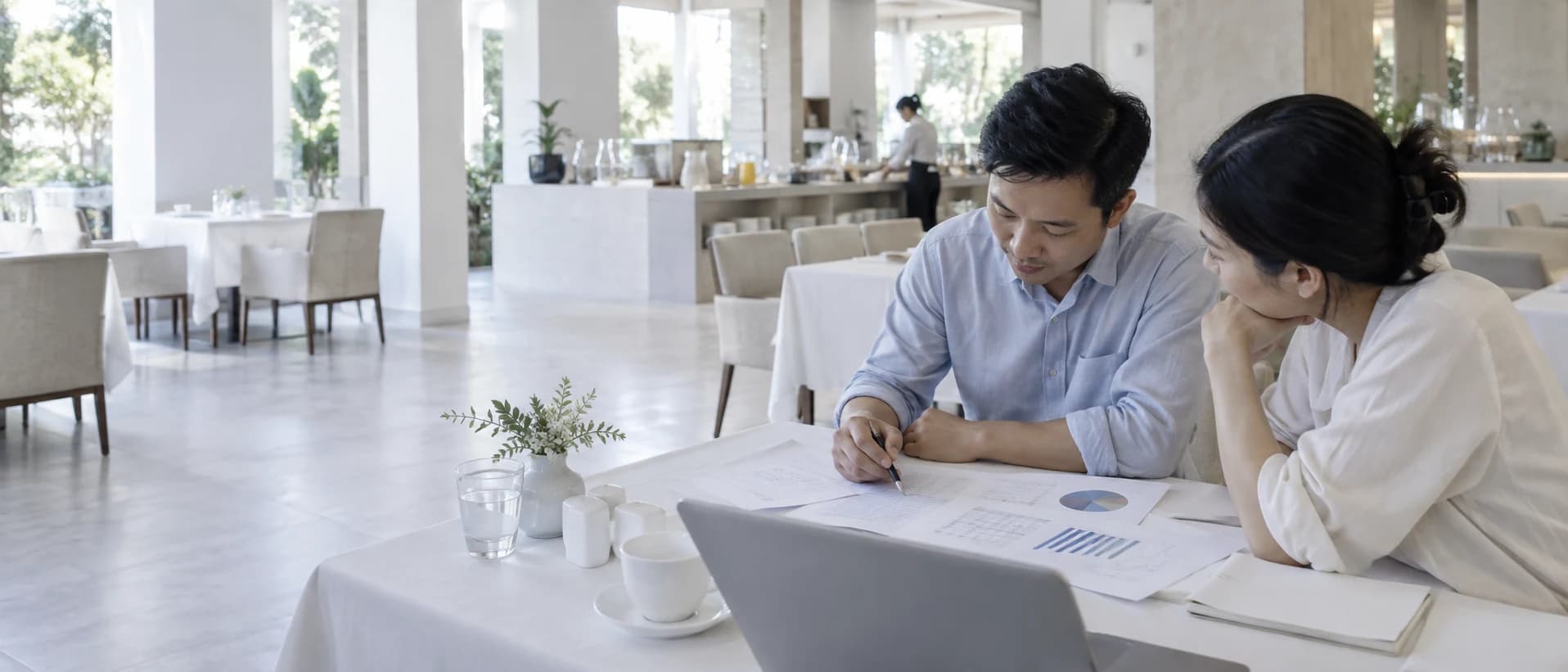 Hai người ngồi cùng một phía bàn trong phòng ăn sáng đã vãn khách, các trang in trải trước mặt.