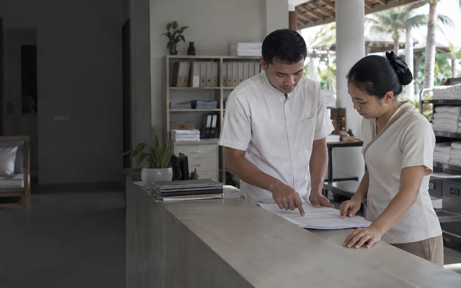 Hai nhân viên khách sạn đứng ở quầy phòng sau, cùng cúi xuống một bản checklist in trên mặt quầy.